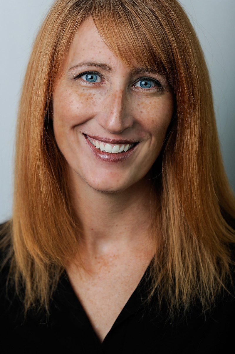 A picture of self-help author, Laura Berg: a white woman with red hair and blue eyes, wearing a black shirt and standing in front of a grey background. 